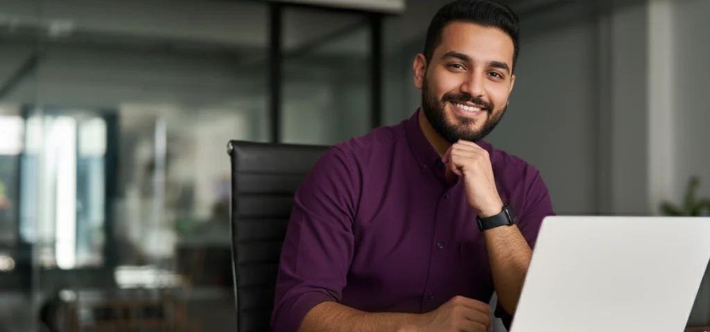 مؤسس شاب يجلس على مكتبه في مكتب حديث، يبتسم للكاميرا وأمامه لابتوب مفتوح.